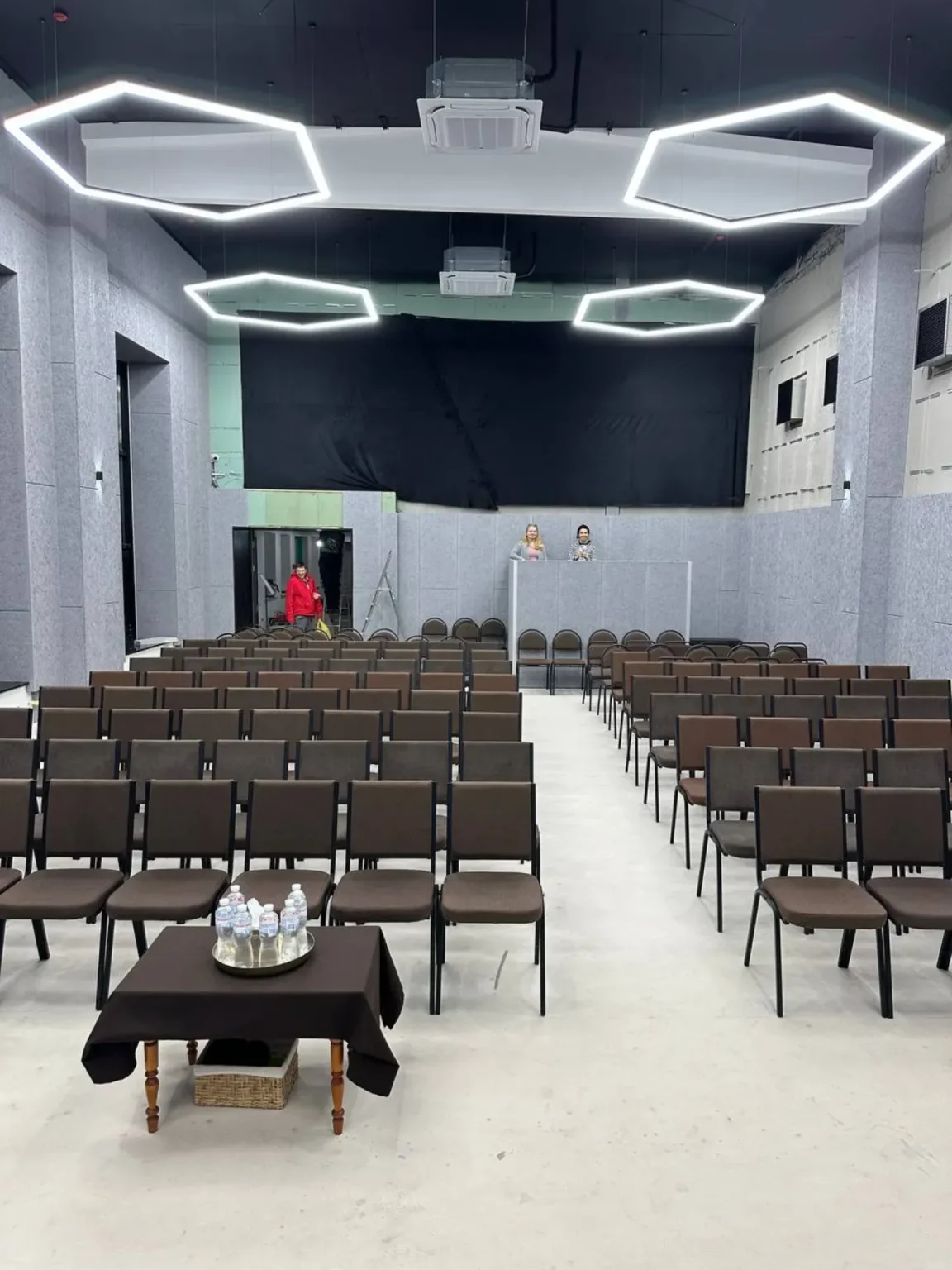 Renovated event or presentation hall featuring modern geometric LED ceiling lights, organized chair layout, and high acoustic walls
