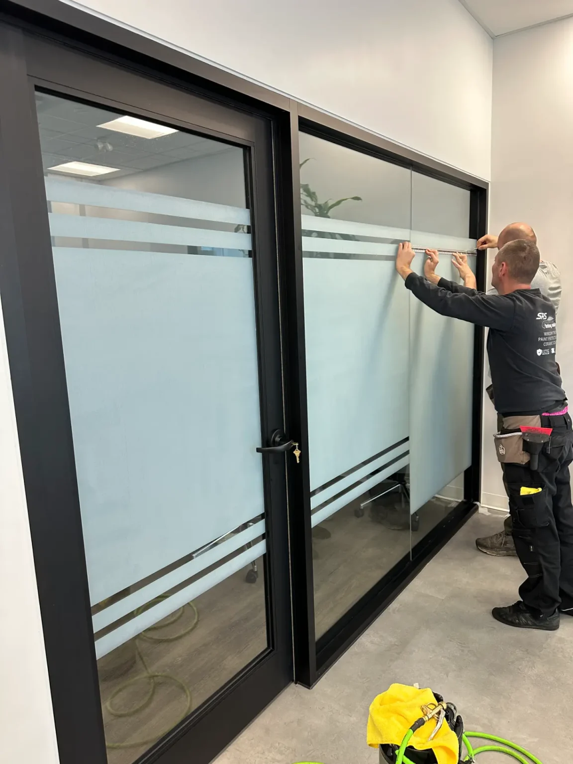 Workers measuring and installing frosted privacy film on a commercial glass office partition with black aluminum framing