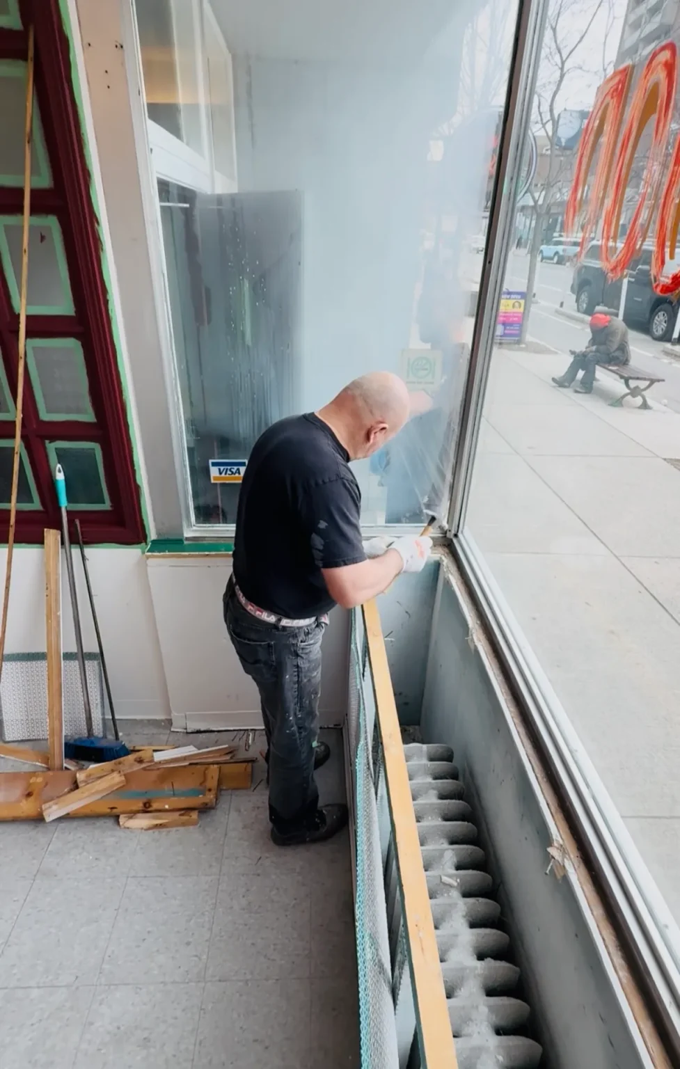 Worker repairing interior storefront window frame near sidewalk-facing commercial display glass with tools and framing wood
