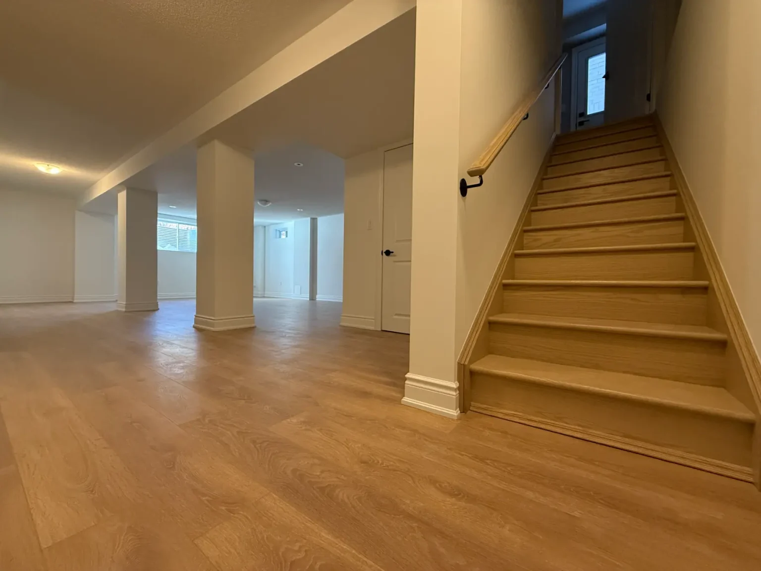 Bright finished basement featuring oak stairs with handrail, wide plank wood-look flooring, and clean white walls creating a warm modern space