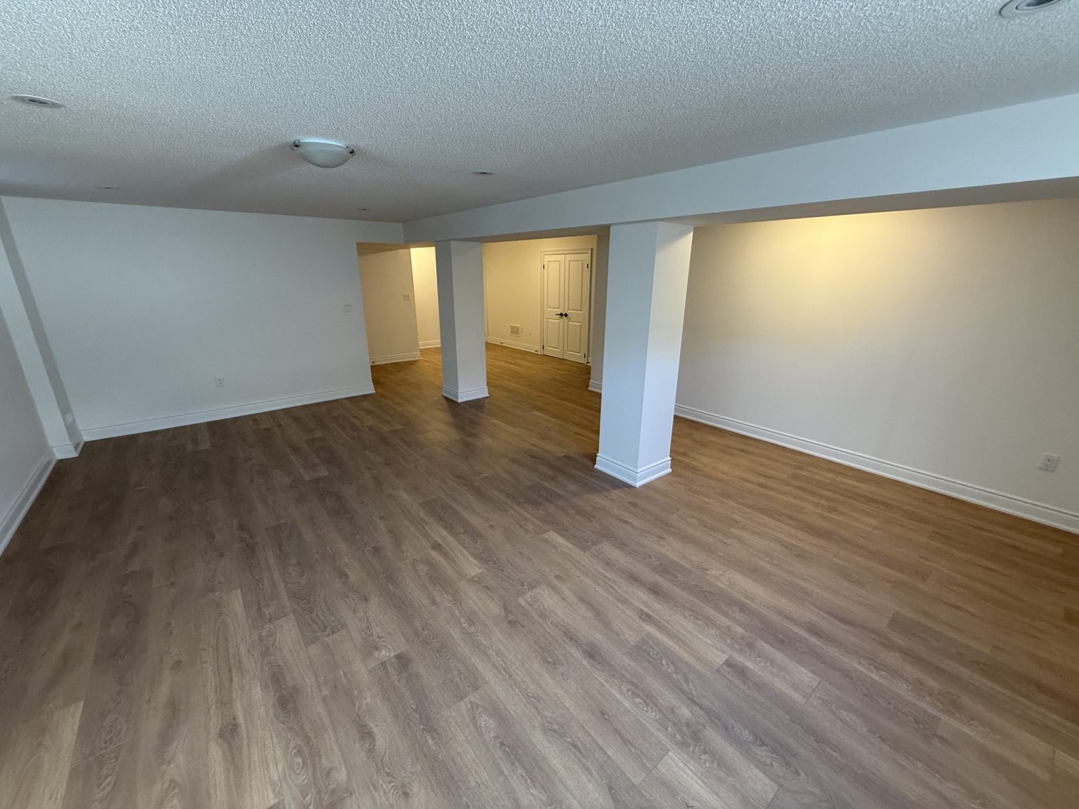 Bright basement renovation featuring warm-toned vinyl flooring, white support beams, recessed ceiling lights, and freshly painted neutral walls