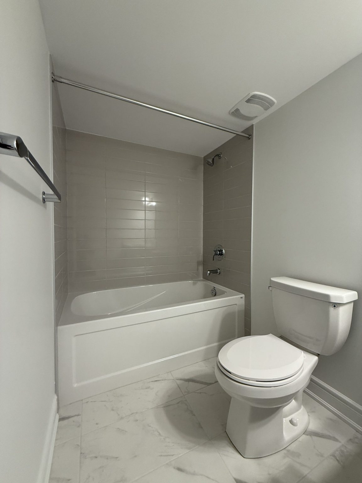 Compact bathroom remodel featuring a built-in soaking tub with grey wall tiles, white toilet, and polished marble-look ceramic floor tiles