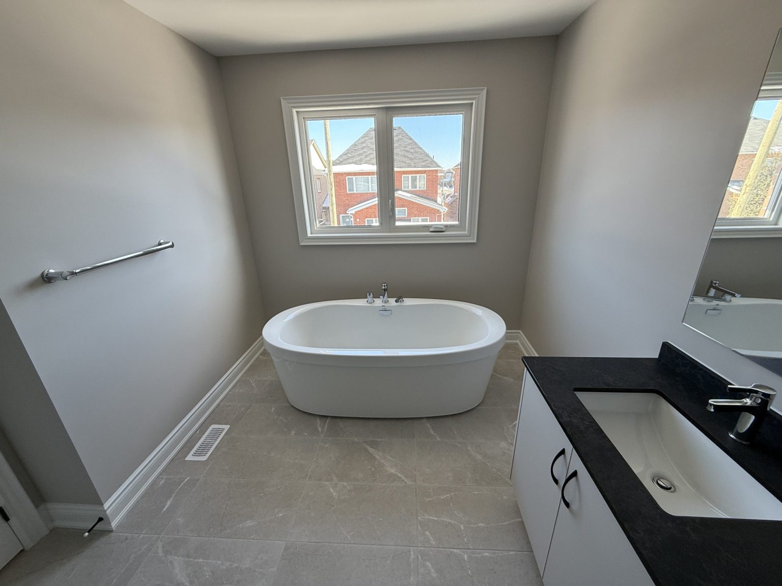 Minimalist bathroom remodel featuring a freestanding soaking tub placed below a bright window, grey tiled flooring, and a sleek white vanity with black countertop