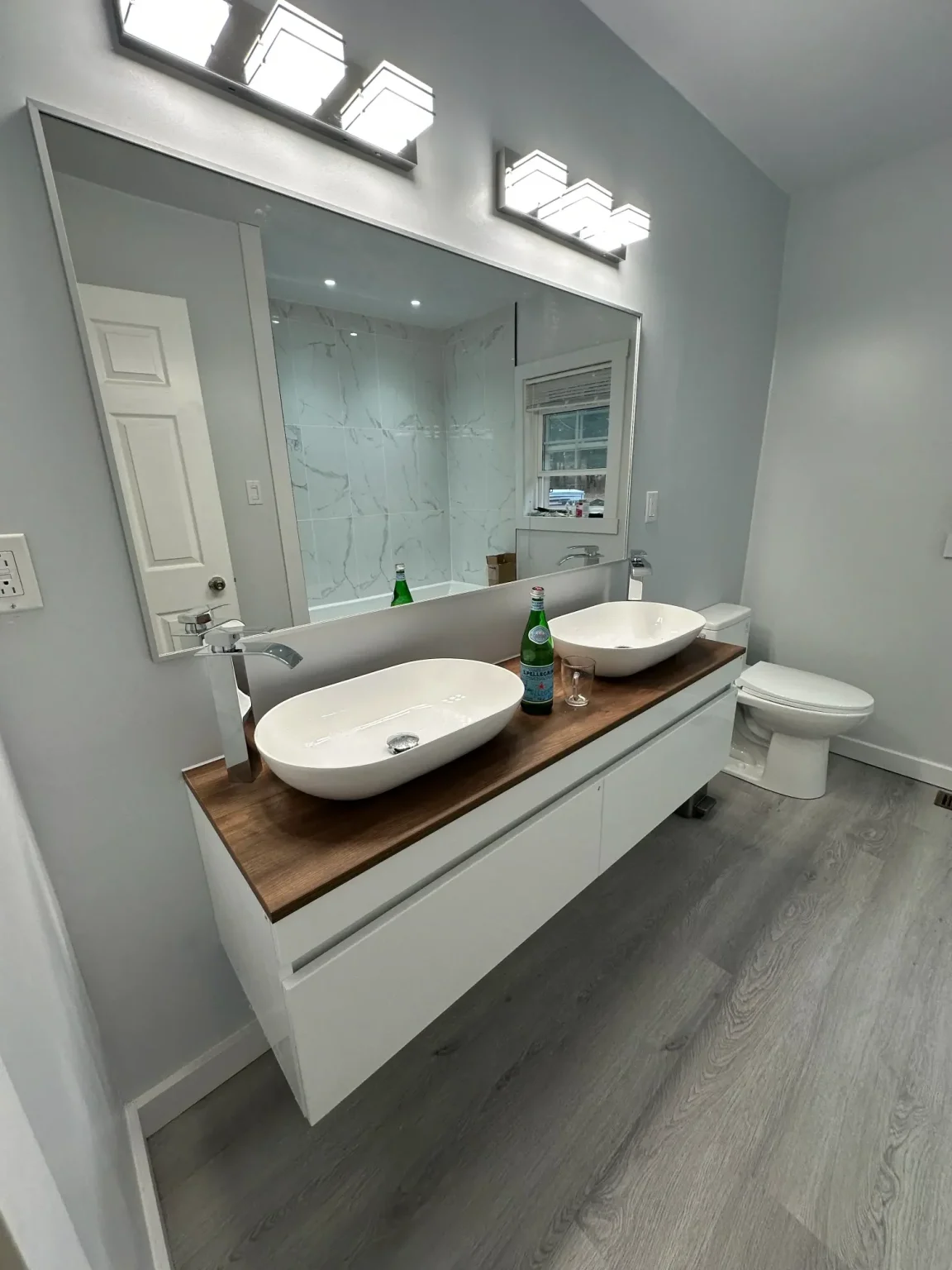 Updated bathroom showcasing double vessel sinks on a wood countertop, sleek LED vanity lights, large wall mirror, and modern marble-style shower wall tiles