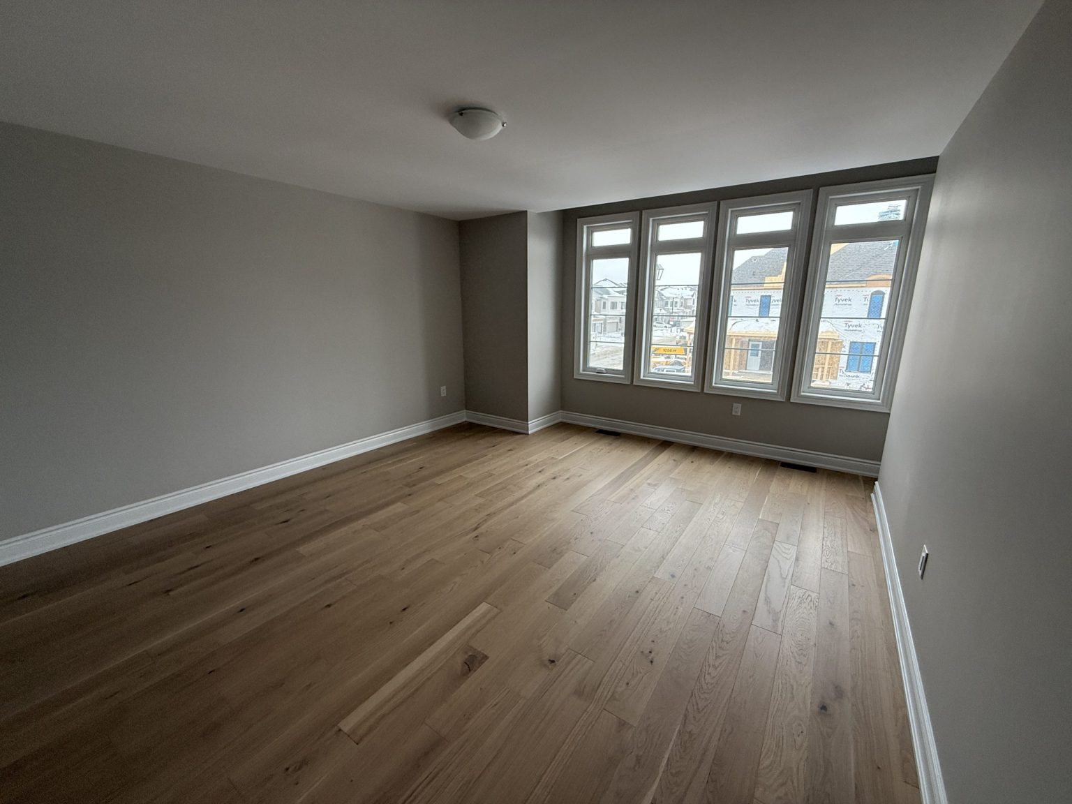 Spacious bedroom renovation featuring new wide-plank hardwood flooring, soft grey walls, and ample natural light from five tall front-facing windows