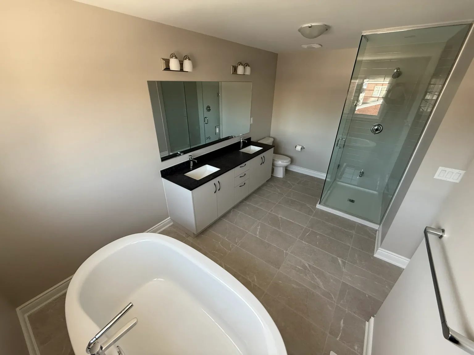 Full bathroom remodel showcasing a freestanding tub, sleek double vanity with black countertops, glass shower enclosure, and soft neutral tile flooring