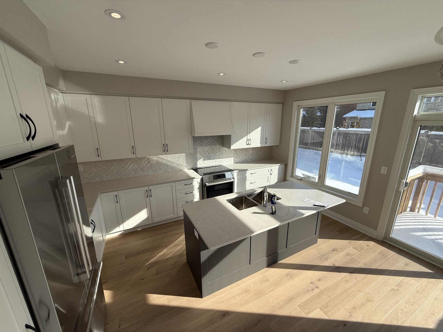 Sunlit kitchen renovation showcasing full-height white cabinets, grey island with sink, quartz counters, and wide-plank oak flooring in a cottage setting