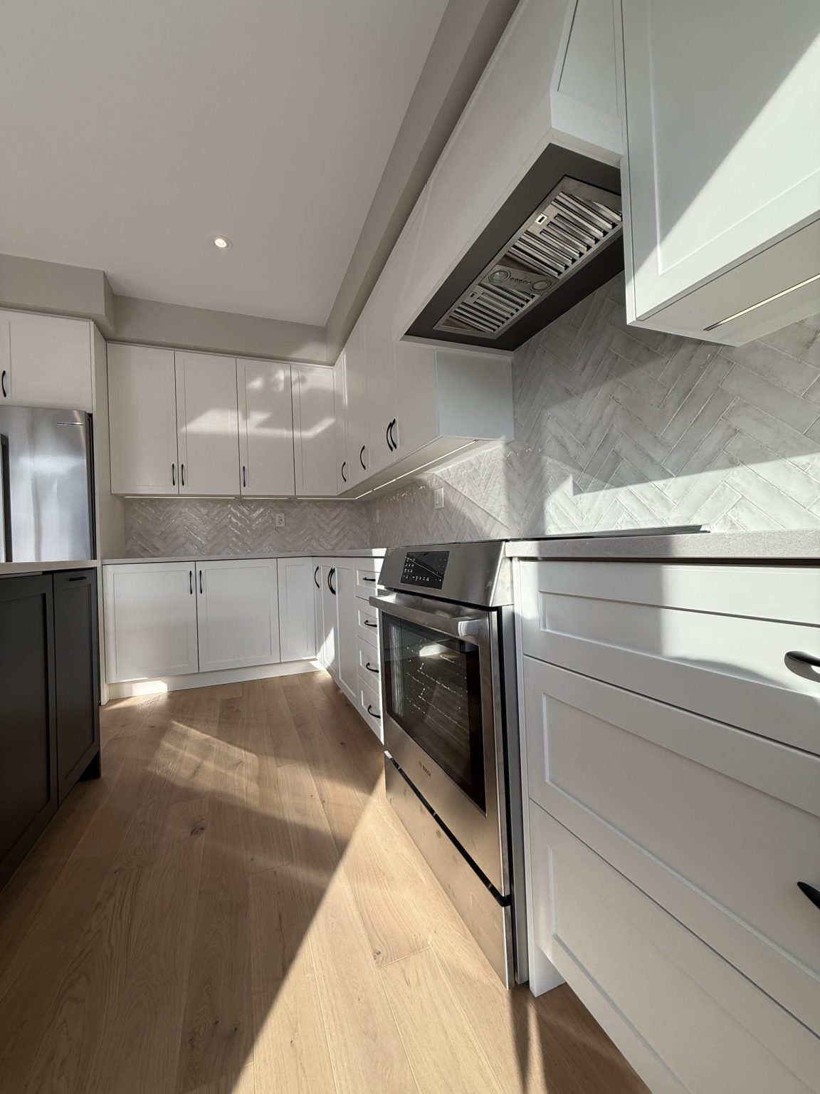 Renovated cottage kitchen showing stainless steel oven, modern vent hood, white cabinetry, and herringbone tile backsplash with warm wood flooring