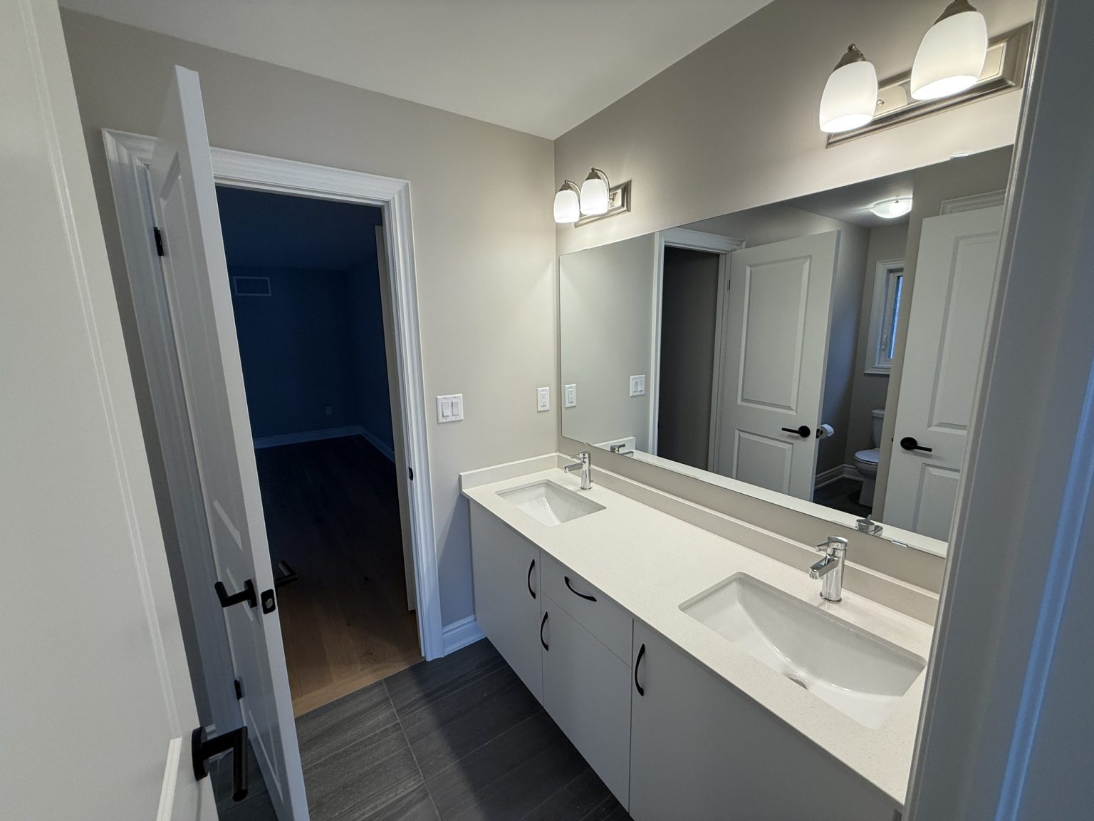 Freshly renovated bathroom featuring a white double sink vanity, sleek chrome faucets, wall-to-wall mirror, and dark floor tiles for contrast