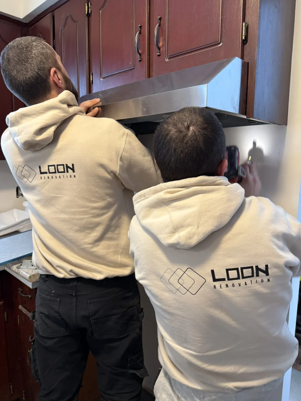 Two Loon Renovation professionals installing a stainless steel range hood under dark wood cabinets during a kitchen upgrade project