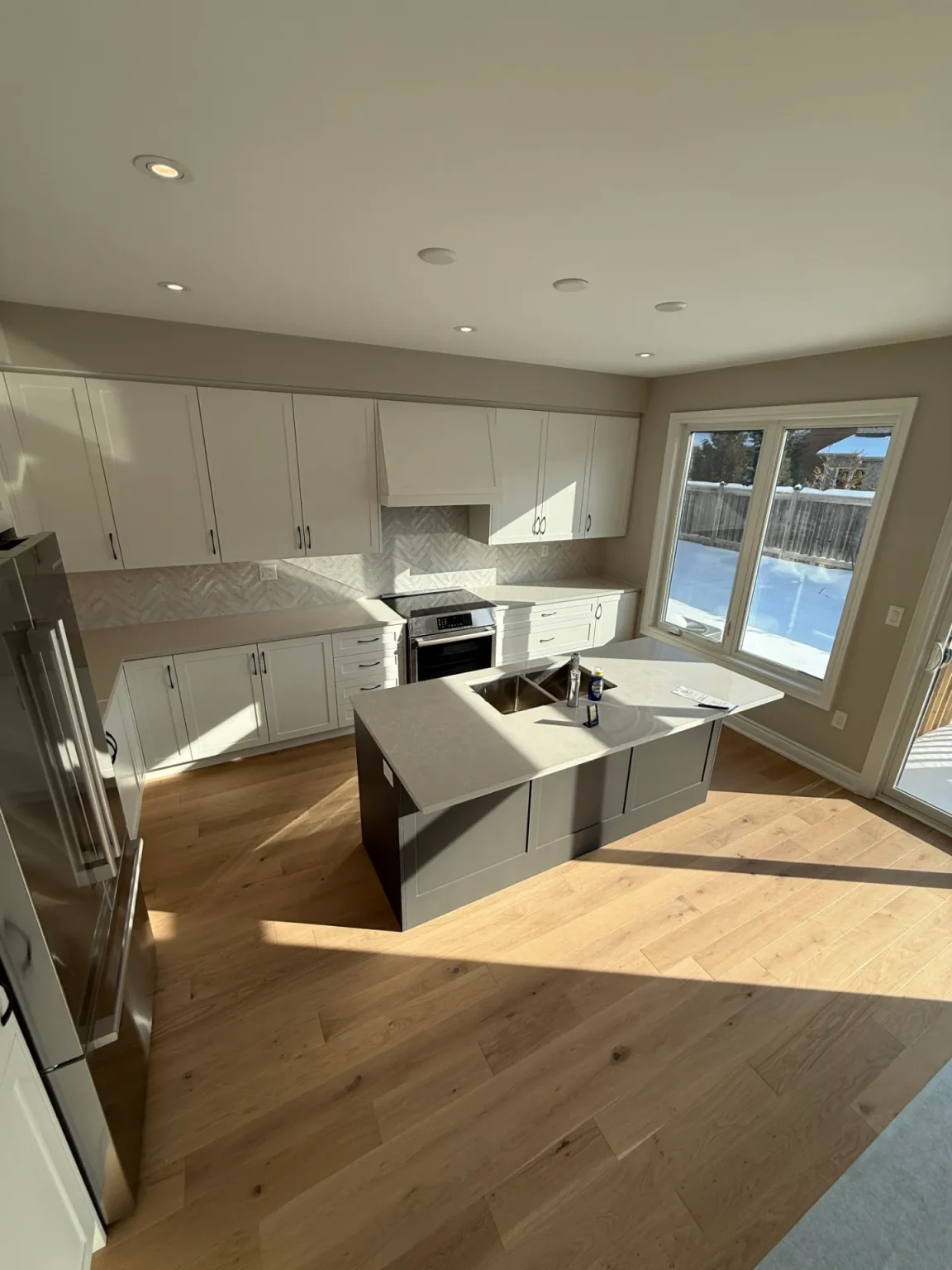 Modern kitchen renovation with shaker-style white cabinets, grey island, natural oak flooring, and large windows by Loon Renovation in Ontario
