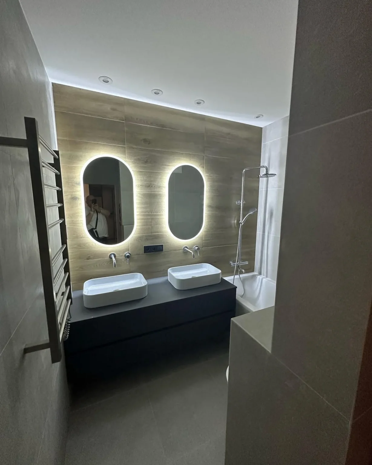 Sleek bathroom renovation with wood accent wall, backlit oval mirrors, vessel sinks, and bathtub-shower combo by Loon Renovation in Ontario