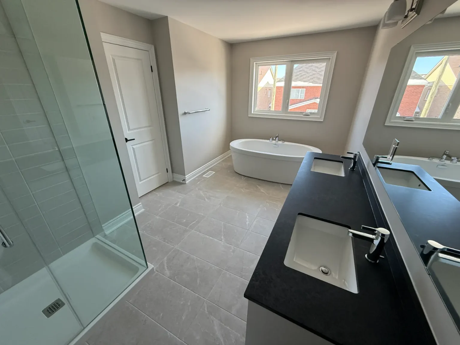 Modern bathroom featuring a freestanding tub under double windows, black countertop double vanity, large glass-enclosed shower, and light grey tile flooring