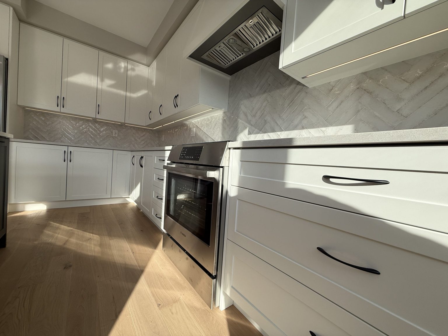 Sleek cottage kitchen renovation featuring white shaker cabinets, under-cabinet lighting, herringbone tile backsplash, and built-in oven with vent hood
