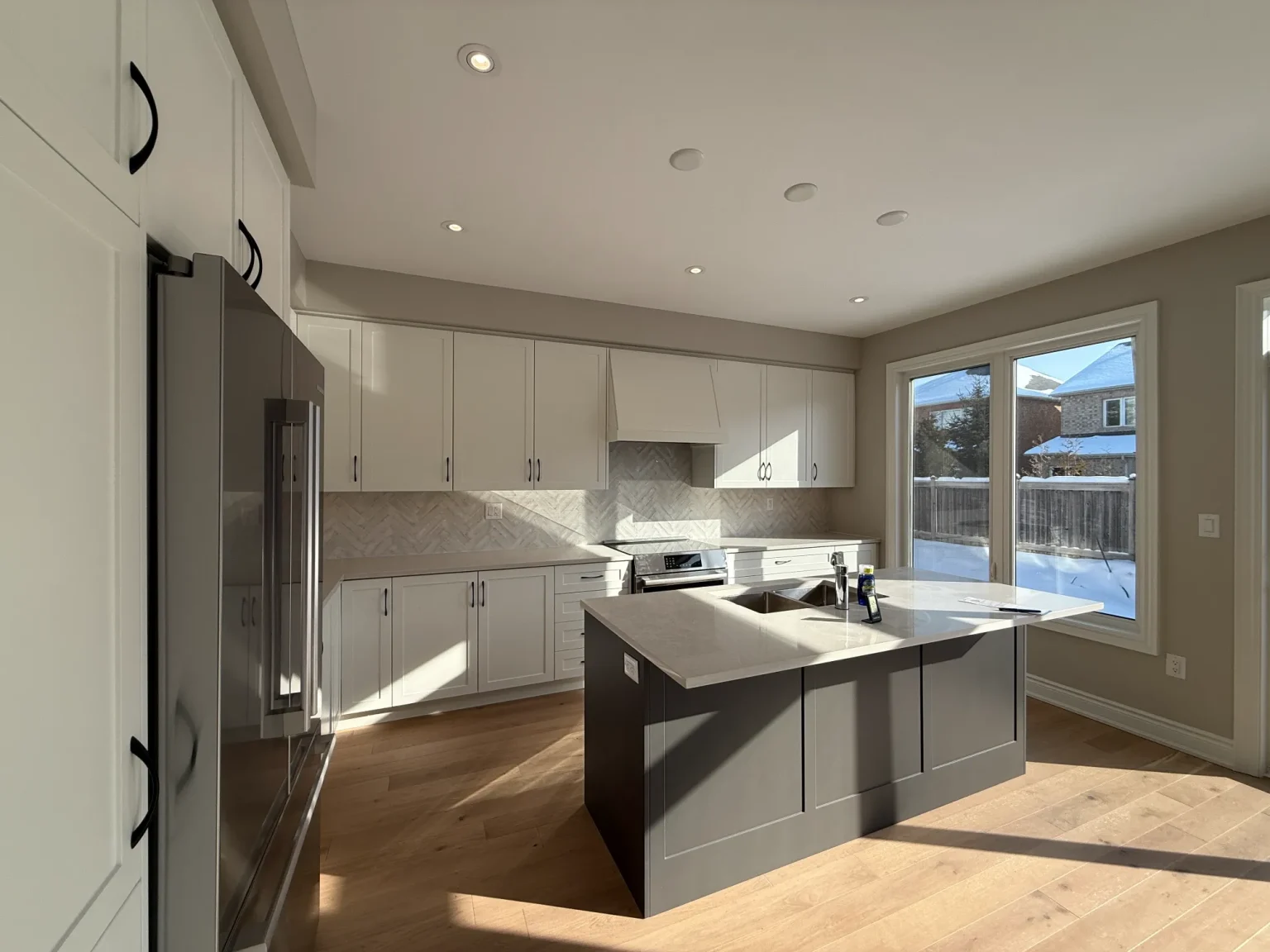 Sunlit cottage kitchen renovation featuring white cabinets, quartz island, and stainless appliances with a view of a snowy backyard through large windows