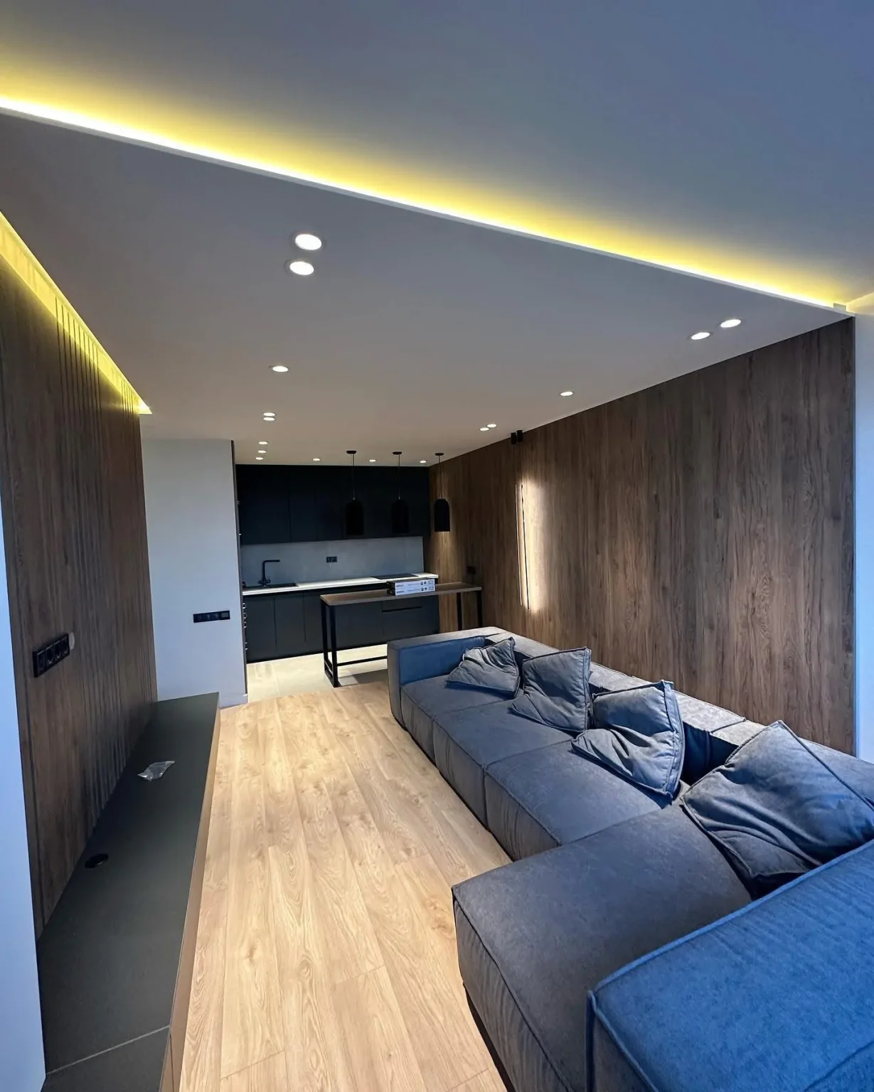 Renovated living space featuring a sleek blue sectional sofa, wood accent walls, recessed LED lighting, and a minimalist kitchen with black finishes