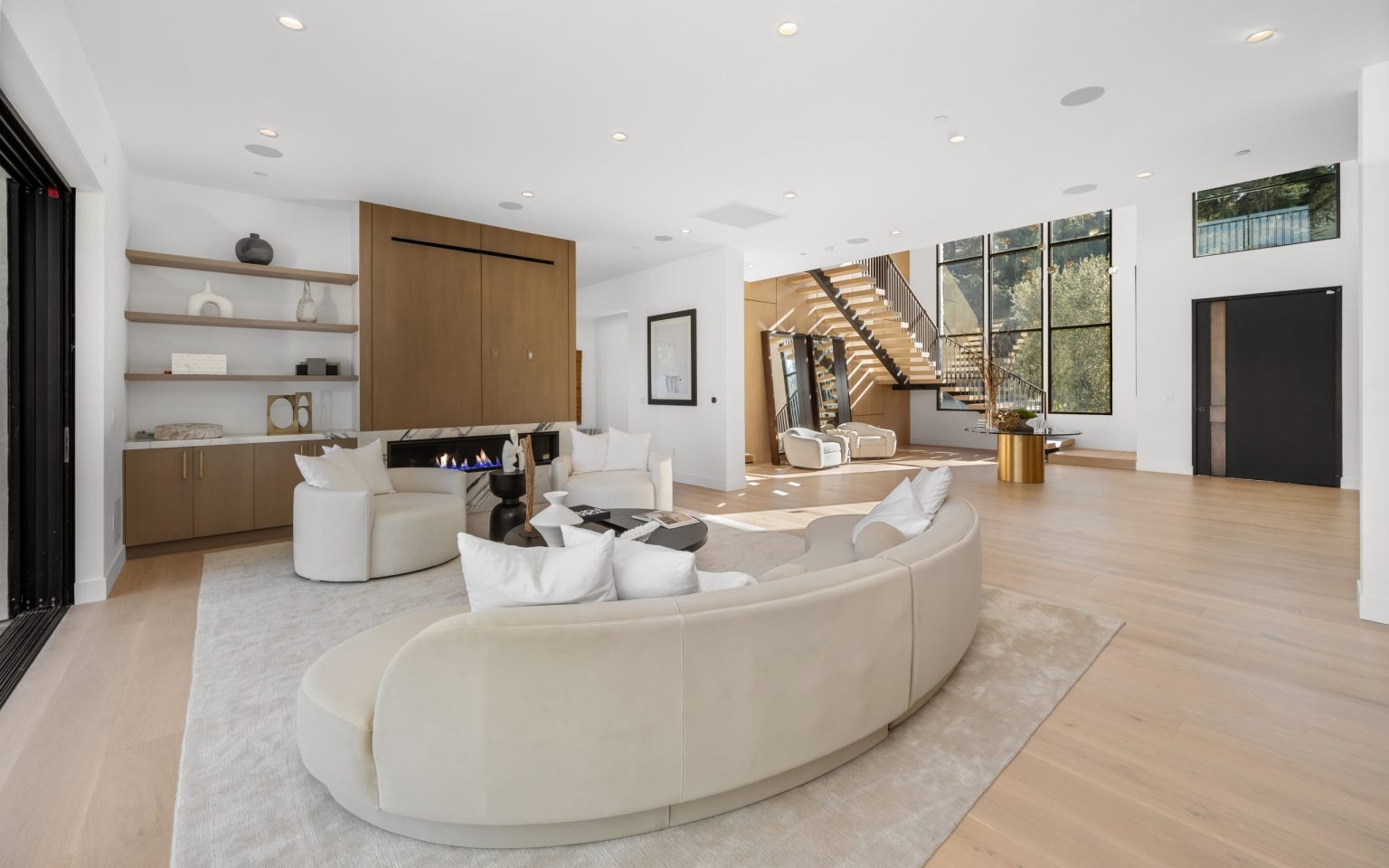 Renovated modern living room featuring white oak flooring, a curved sectional, built-in shelving, and a contemporary gas fireplace with wood paneling