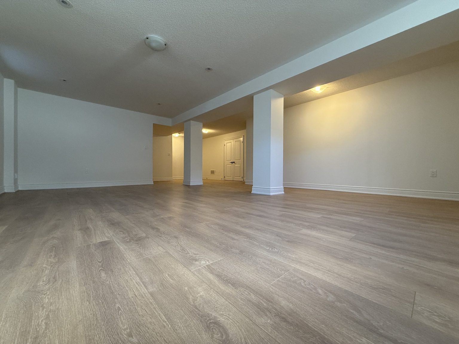 Spacious finished basement with light-toned wide plank flooring, clean white walls, recessed lighting, and support columns for an open layout design