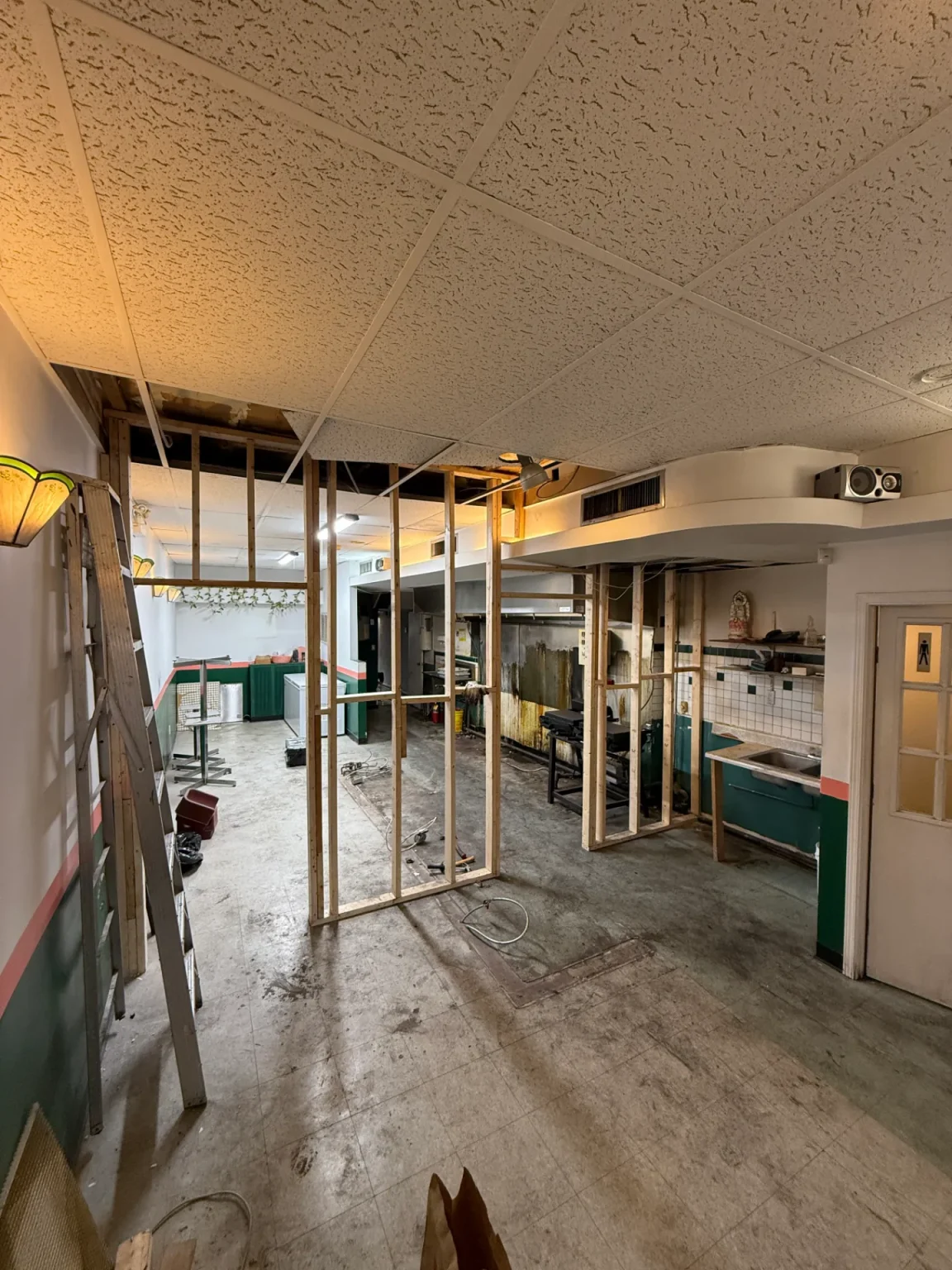 Early stage of restaurant renovation with partial wall framing, exposed ceiling tiles, and outdated kitchen equipment pre-demolition