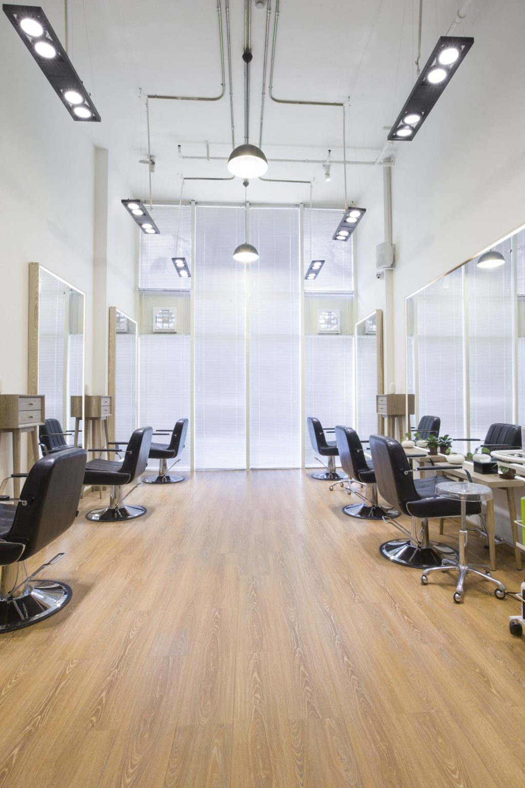 Spacious salon interior featuring wooden flooring, black styling chairs, full-length mirrors, and high ceilings with modern lighting