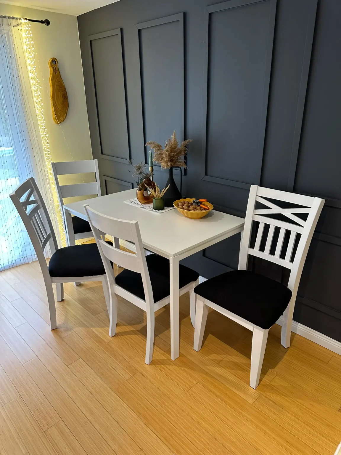 Compact dining area featuring white table and chairs with black cushions, warm wood floors, and a dark panel accent wall for modern contrast